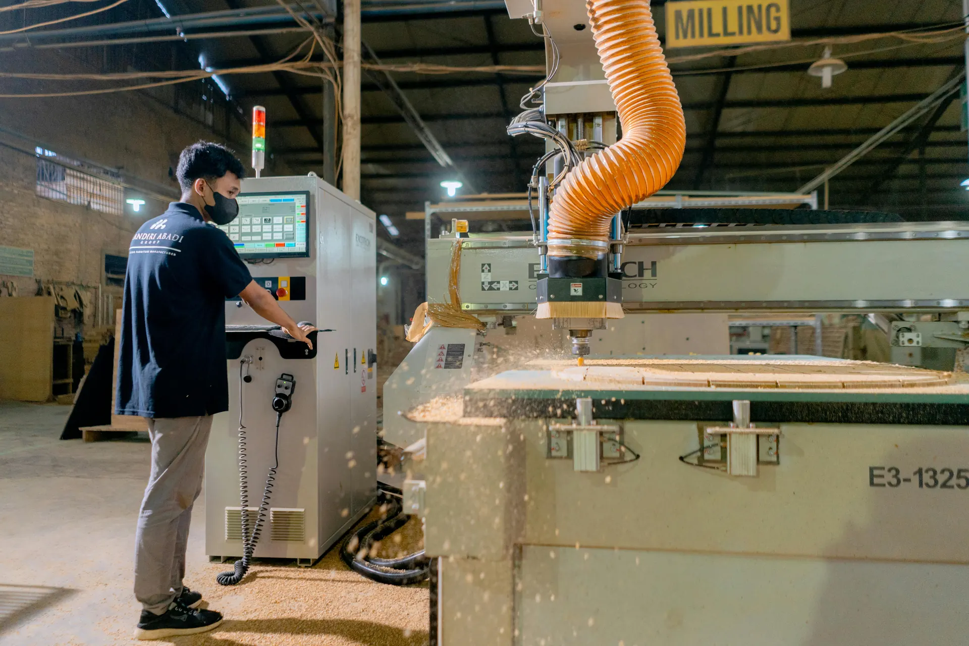 A CNC router cutting a wood panel on a shop floor, sawdust in the air, an operator at the control desk
